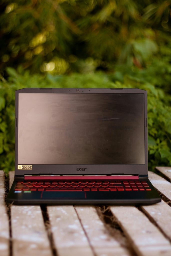 A gaming laptop with red keyboard on a wooden surface outdoors, surrounded by greenery.